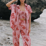 A woman stands on a beach wearing a matching pink floral lounge set with a short-sleeve button-up shirt, cropped tank top, and wide-leg pants. Large dark rocks and shallow water are behind her.