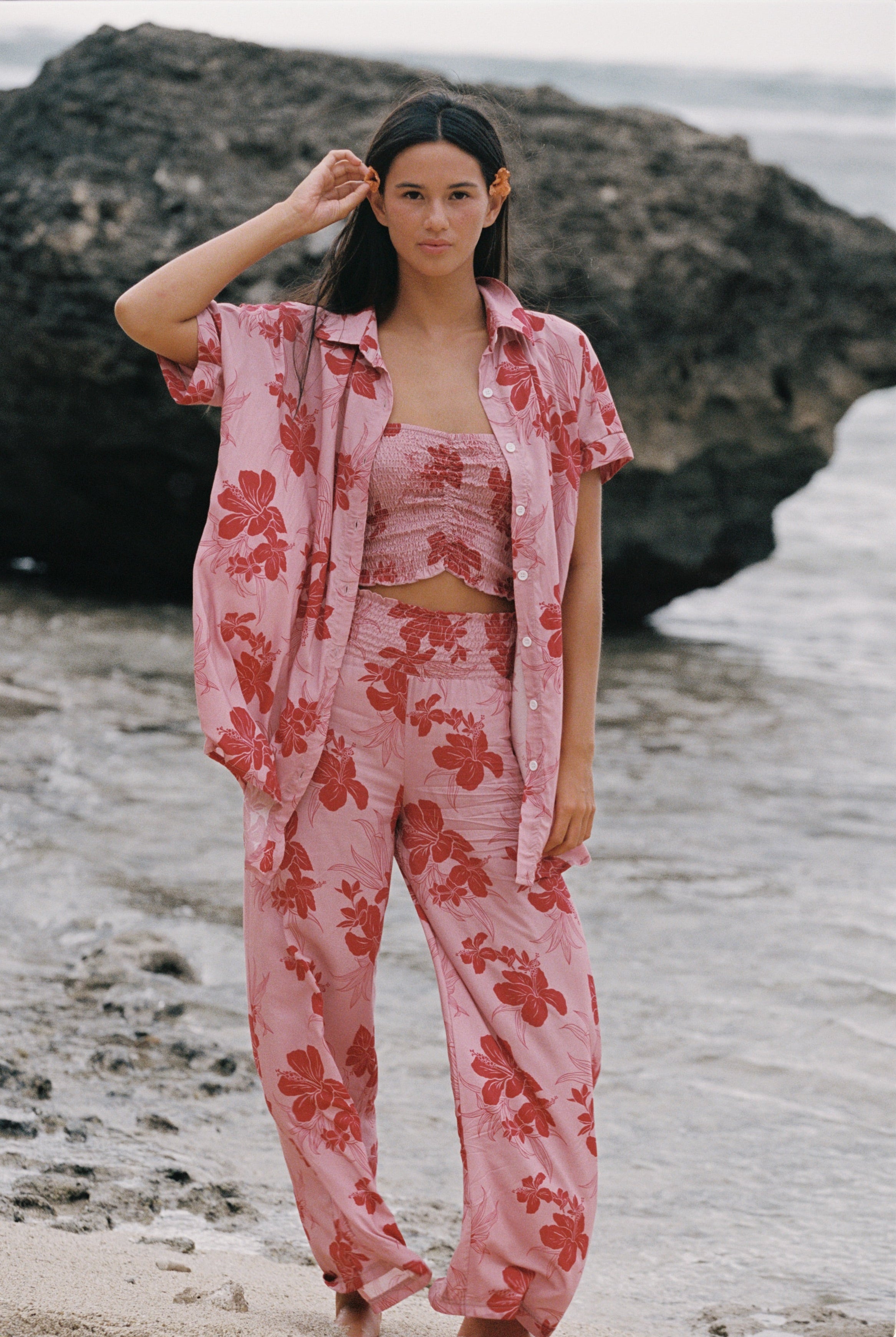 A woman stands on a beach wearing a matching pink floral lounge set with a short-sleeve button-up shirt, cropped tank top, and wide-leg pants. Large dark rocks and shallow water are behind her.