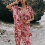 A woman stands on a beach wearing a pink two-piece pajama set with a red floral print, consisting of a cropped tank top and matching pants, with an unbuttoned shirt jacket draped off her shoulders.