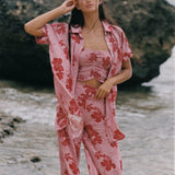 A woman stands on a beach wearing a pink two-piece pajama set with a red floral print, consisting of a cropped tank top and matching pants, with an unbuttoned shirt jacket draped off her shoulders.