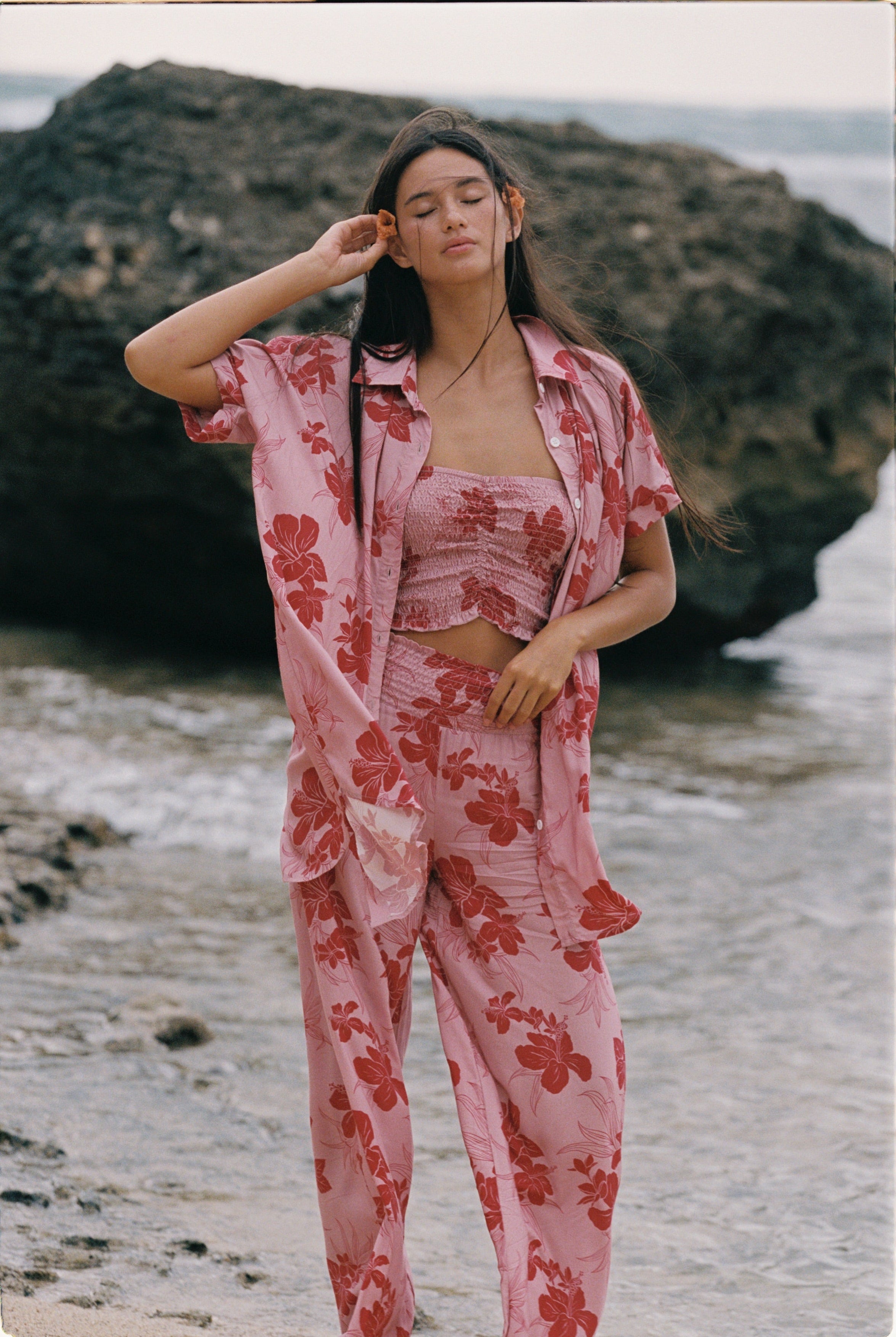 A woman stands on a beach wearing a pink two-piece pajama set with a red floral print, consisting of a cropped tank top and matching pants, with an unbuttoned shirt jacket draped off her shoulders.