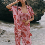 A woman stands on a beach wearing a pink two-piece pajama set with a red floral print, consisting of a cropped tank top and matching pants, with an unbuttoned shirt jacket draped off her shoulders.
