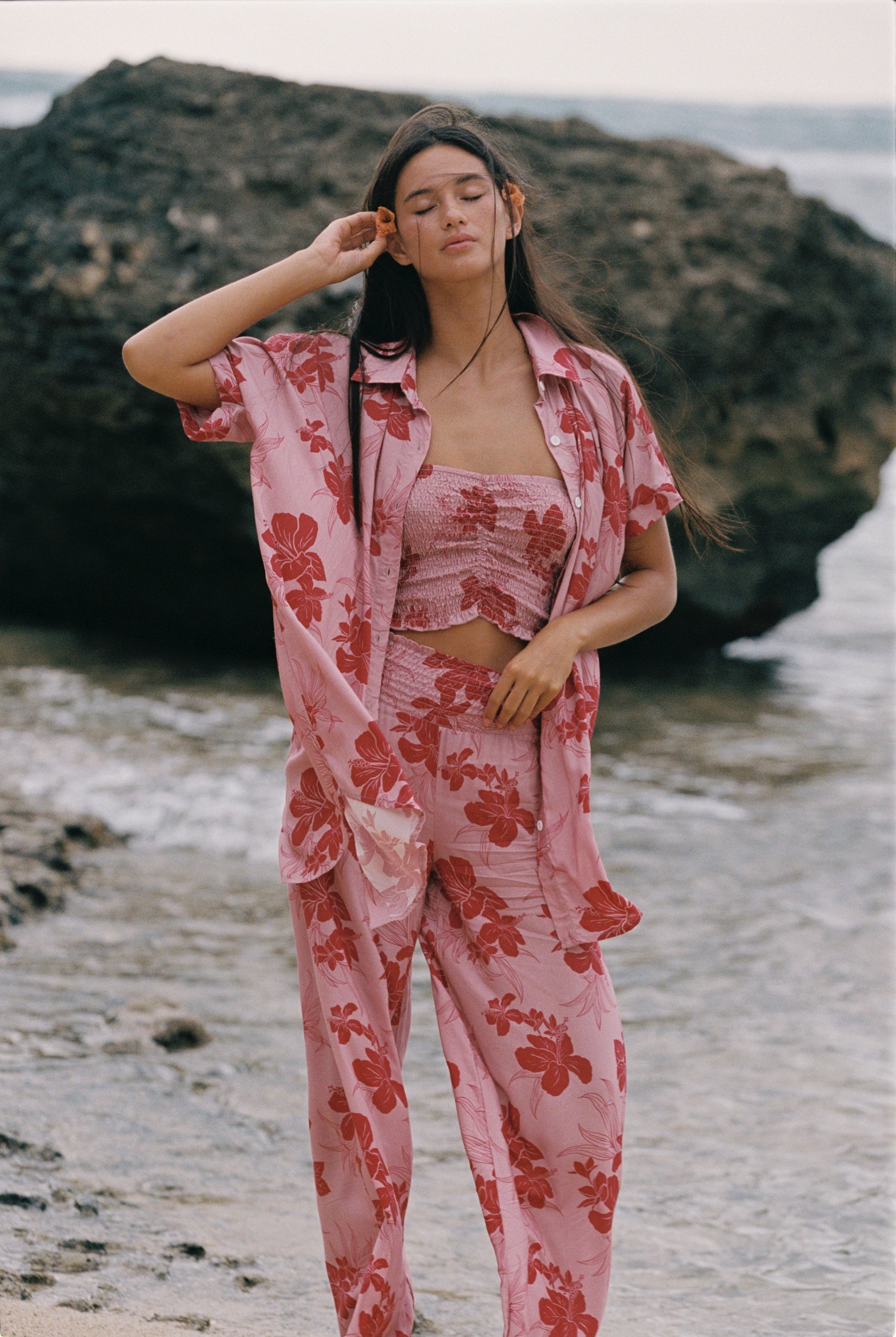 A woman stands on a beach wearing a pink two-piece pajama set with a red floral print, consisting of a cropped tank top and matching pants, with an unbuttoned shirt jacket draped off her shoulders.