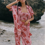 A woman stands on a beach wearing a pink two-piece pajama set with a red floral print, consisting of a cropped tank top and matching pants, with an unbuttoned shirt jacket draped off her shoulders.