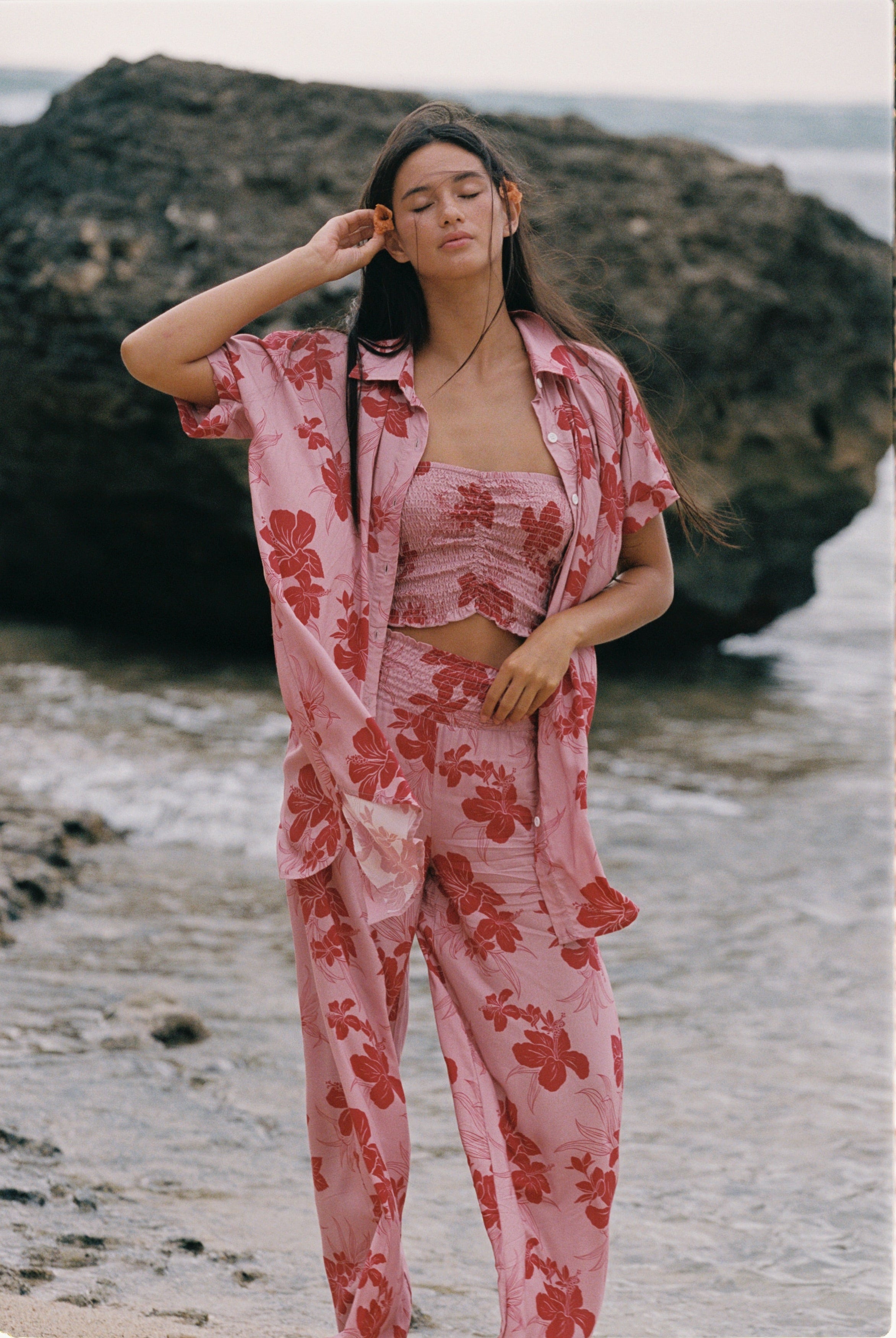 A woman stands on a beach wearing a pink two-piece pajama set with a red floral print, consisting of a cropped tank top and matching pants, with an unbuttoned shirt jacket draped off her shoulders.