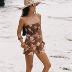 A woman stands on a sandy beach wearing a brown floral strapless tube top and matching shorts, with a wide-brimmed straw hat in the background.