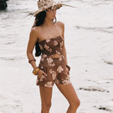 A woman stands on a sandy beach wearing a brown floral strapless tube top and matching shorts, with a wide-brimmed straw hat in the background.
