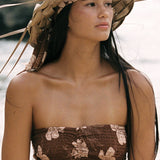 A woman kneeling on a sandy beach wears a brown floral strapless top and matching bottoms, with a large woven headpiece and bracelets. Ocean waves and dark rocks are visible in the background.