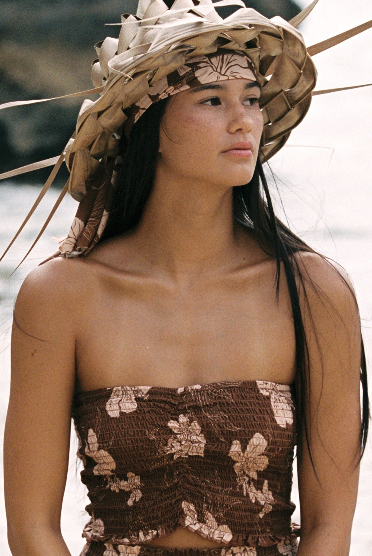 A woman kneeling on a sandy beach wears a brown floral strapless top and matching bottoms, with a large woven headpiece and bracelets. Ocean waves and dark rocks are visible in the background.