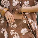 A close-up of a person wearing a matching brown floral lounge set with a button-front shirt and shorts, styled with wooden bangles.