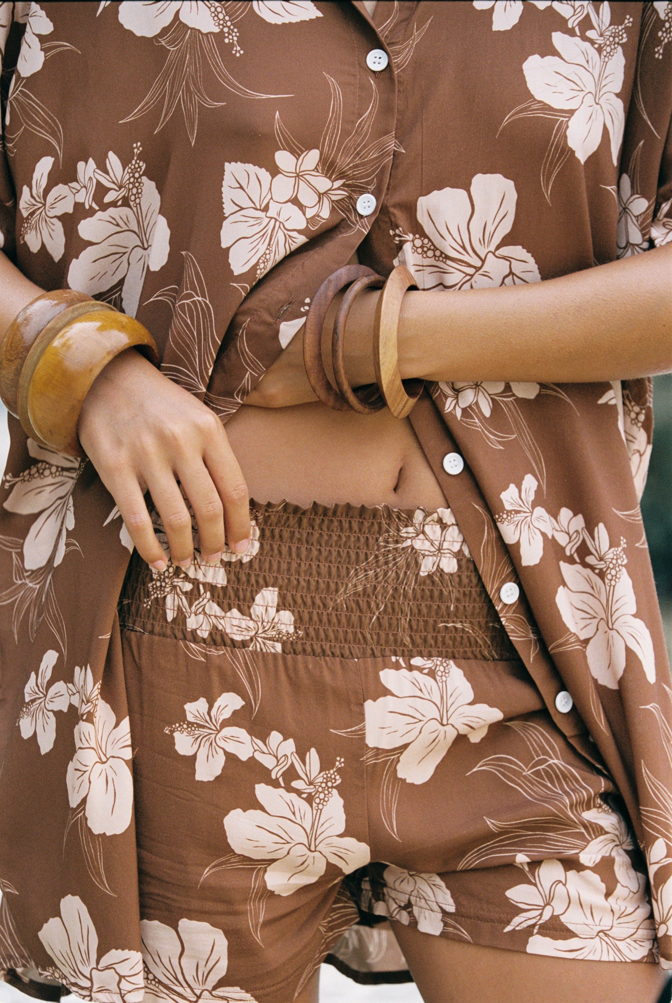A close-up of a person wearing a matching brown floral lounge set with a button-front shirt and shorts, styled with wooden bangles.